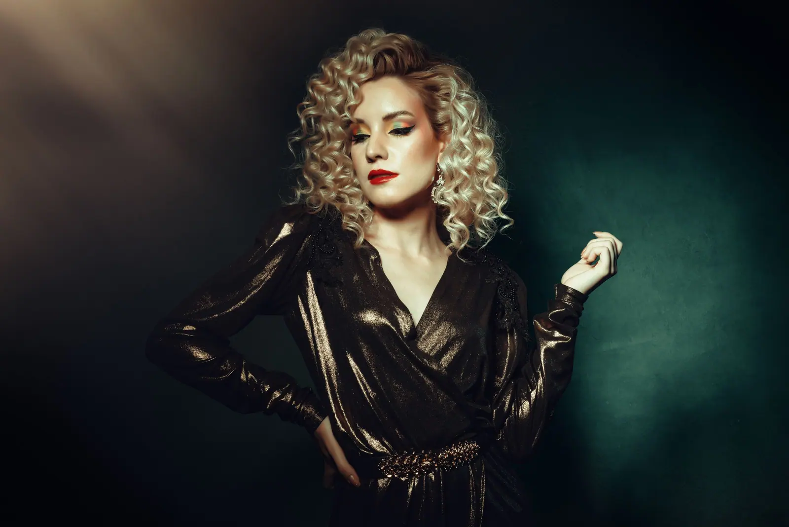 Woman with voluminous blonde curls in a dark satin dress against a deep teal backdrop