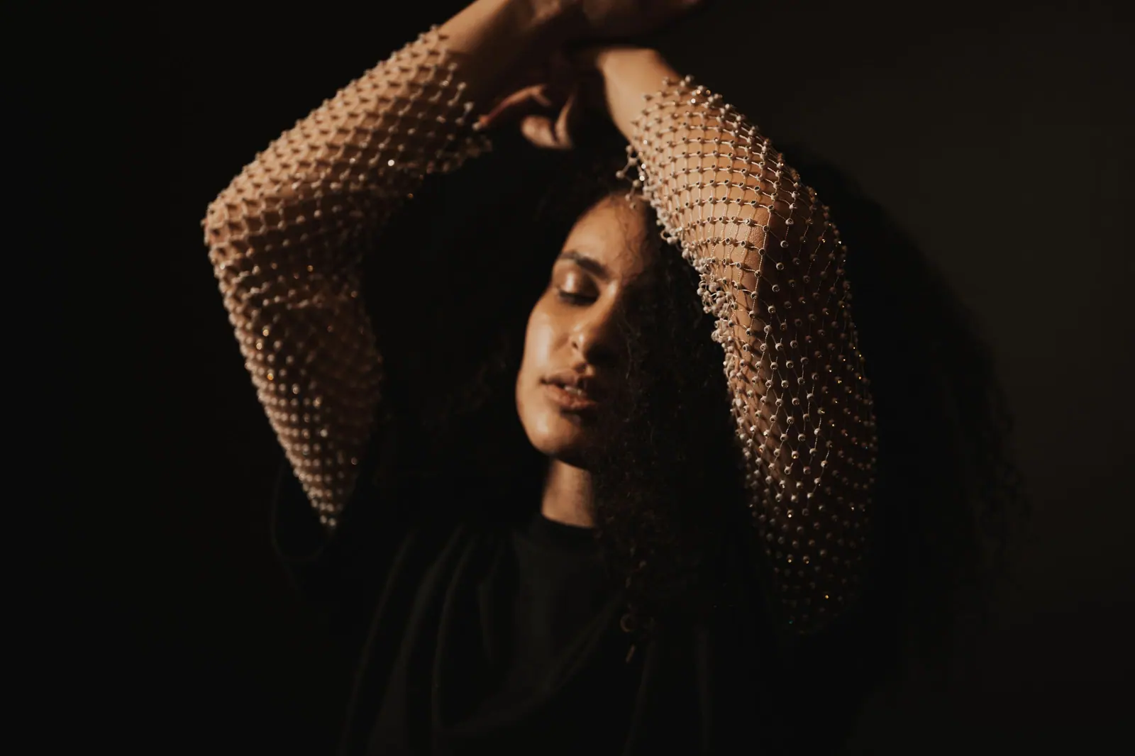 Woman with long dark curls lifting her knit sleeves overhead in low, warm light