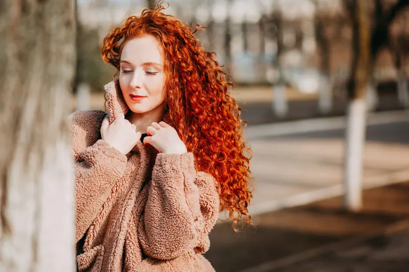 A redhead woman in a fluffy coat enjoys a sunny fall day, showcasing natural beauty and vibrant hair.