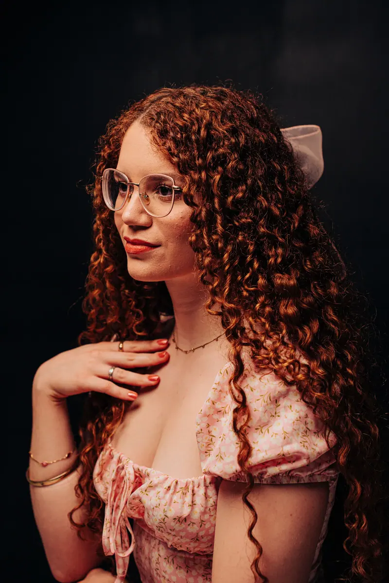 A woman with curly red hair wearing glasses poses elegantly against a dark background.