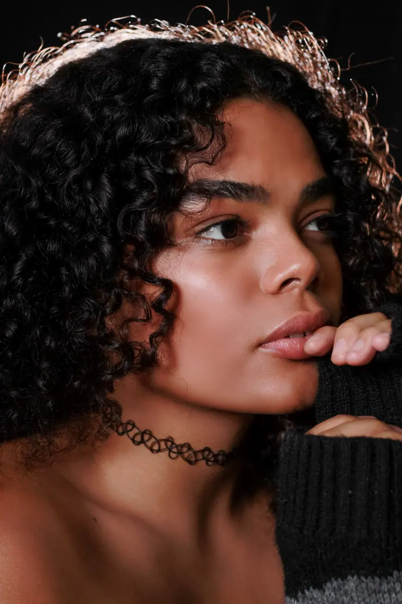 Close-up portrait of a woman with curly hair, wearing a choker necklace, gazing thoughtfully.