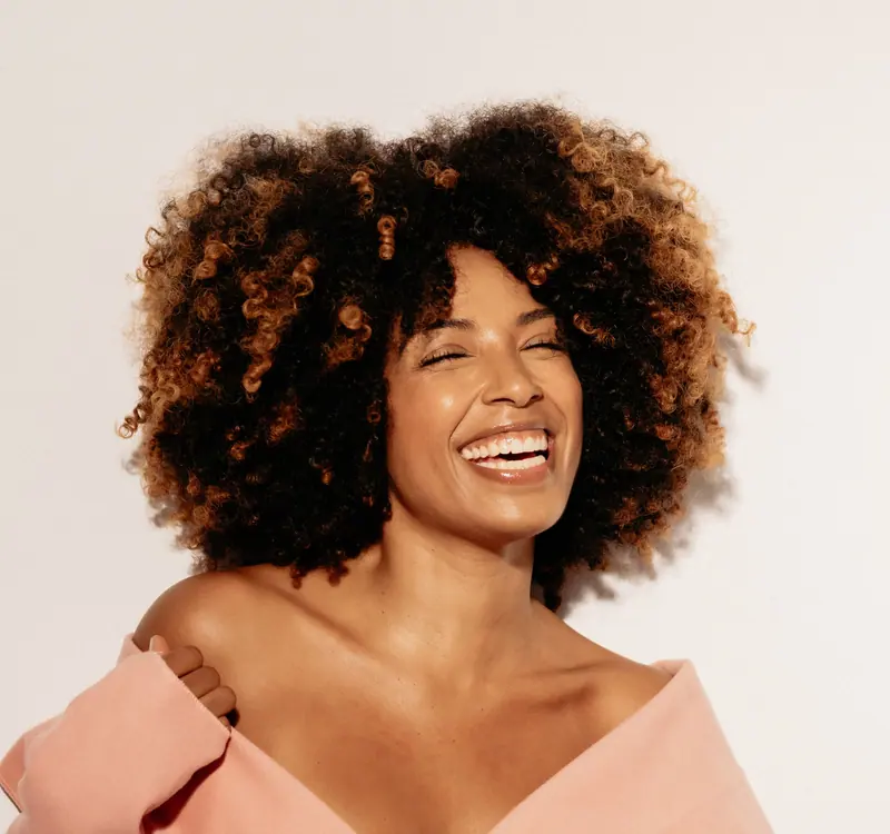 A cheerful woman with curly hair smiling brightly, wearing an off-shoulder top.