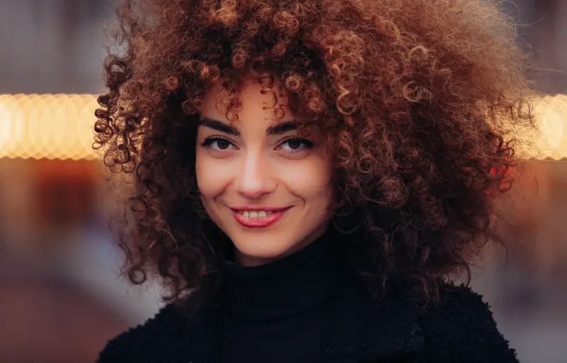 A stunning portrait of a woman smiling warmly, featuring natural curly hair and a stylish black outfit.