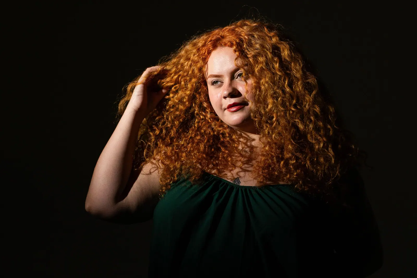 Woman with voluminous copper curls in a dark green dress, lit softly against black