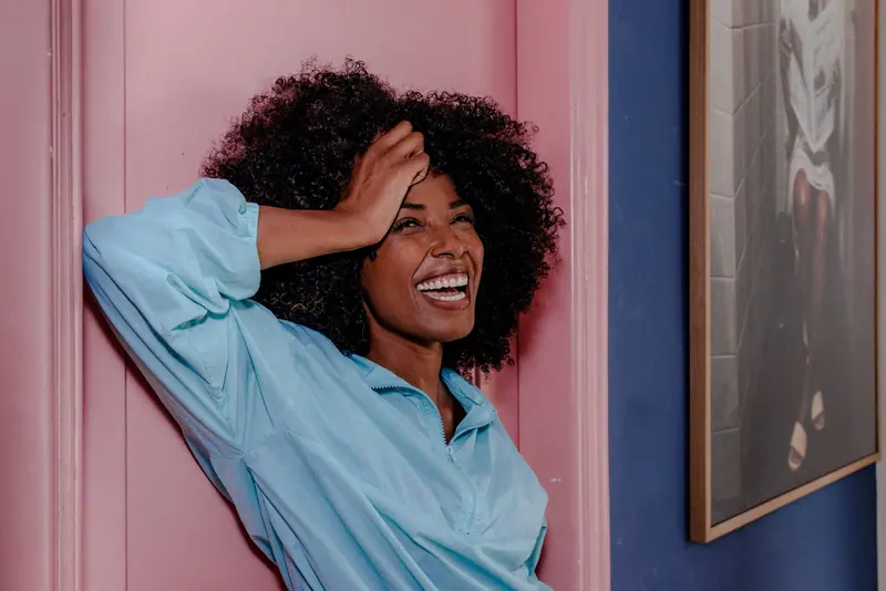 Smiling woman in blue shirt laughing and leaning against a pink and blue wall, exuding joy.