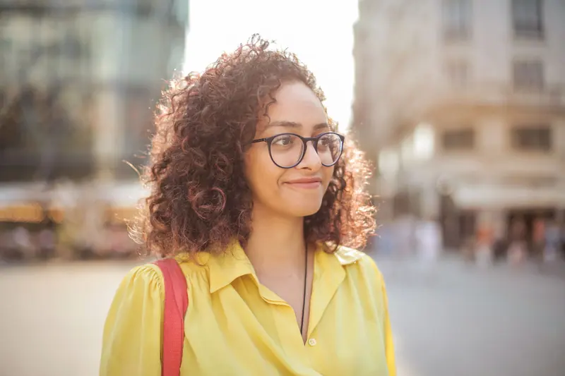Portrait of a smiling woman with curly hair and glasses in an urban setting.