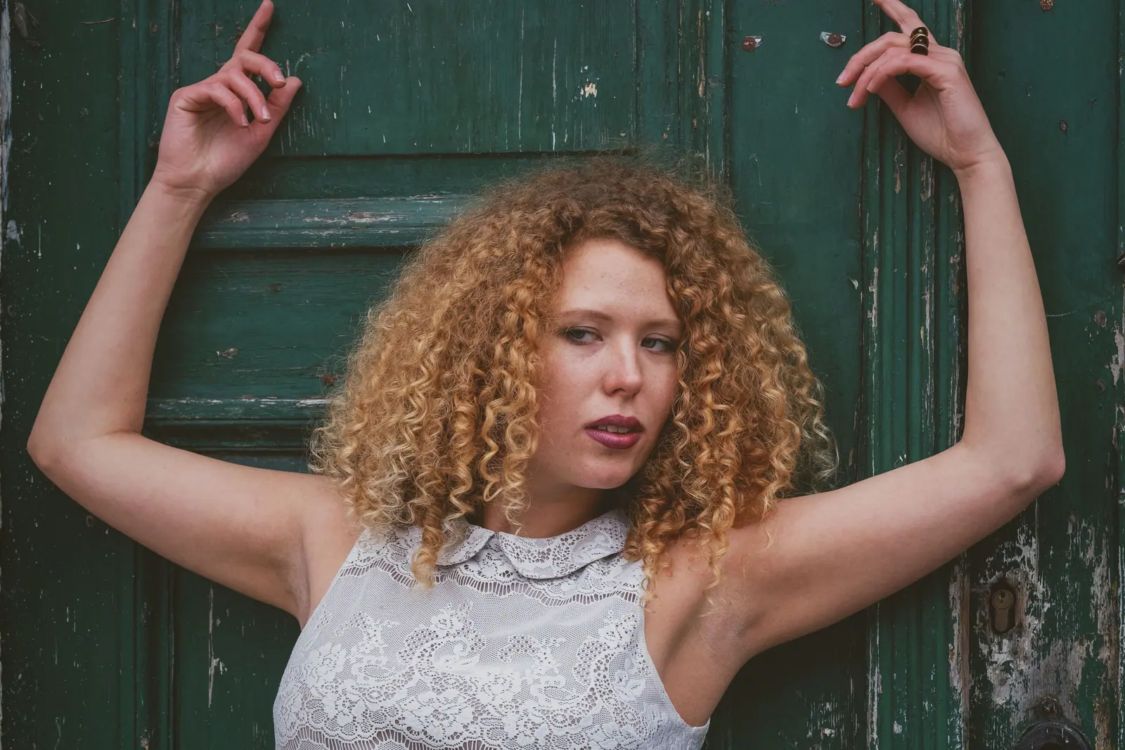Woman with golden spiral curls raising her arms against a weathered green door