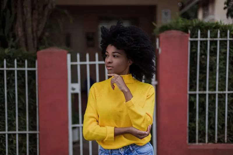 Black woman in a yellow sweater stands outdoors, lost in thought. Tranquil, thoughtful vibe.