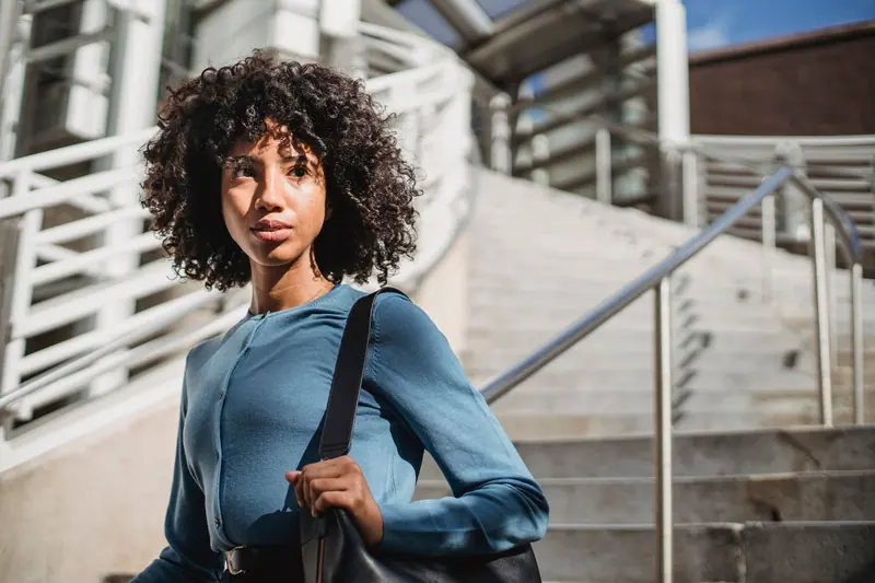 A fashionable woman with curly hair and a bag stands thoughtfully on city stairs.