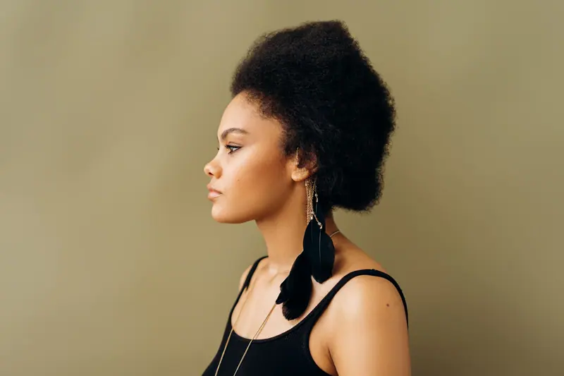 Side profile portrait of a woman with natural afro hairstyle and feather earrings against a brown background.