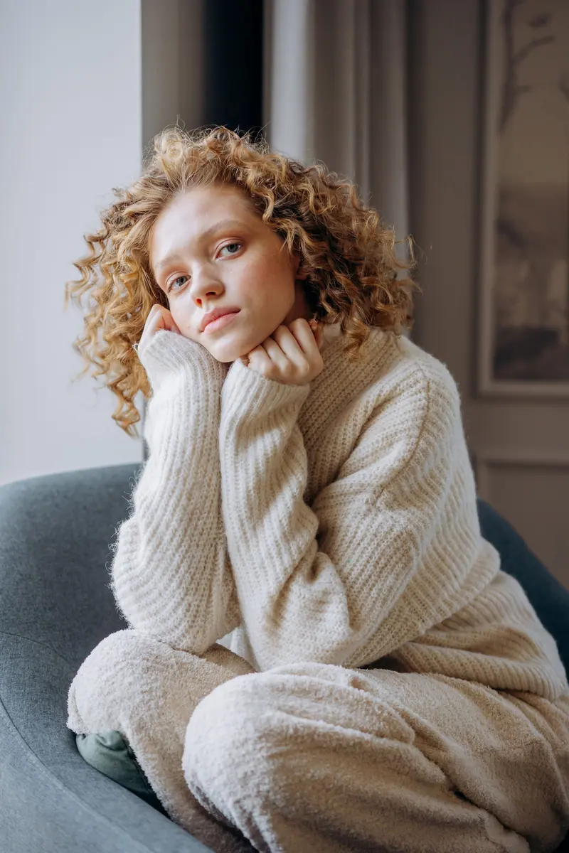 Young woman in a cozy sweater sits comfortably on a sofa indoors, exuding warmth and relaxation.