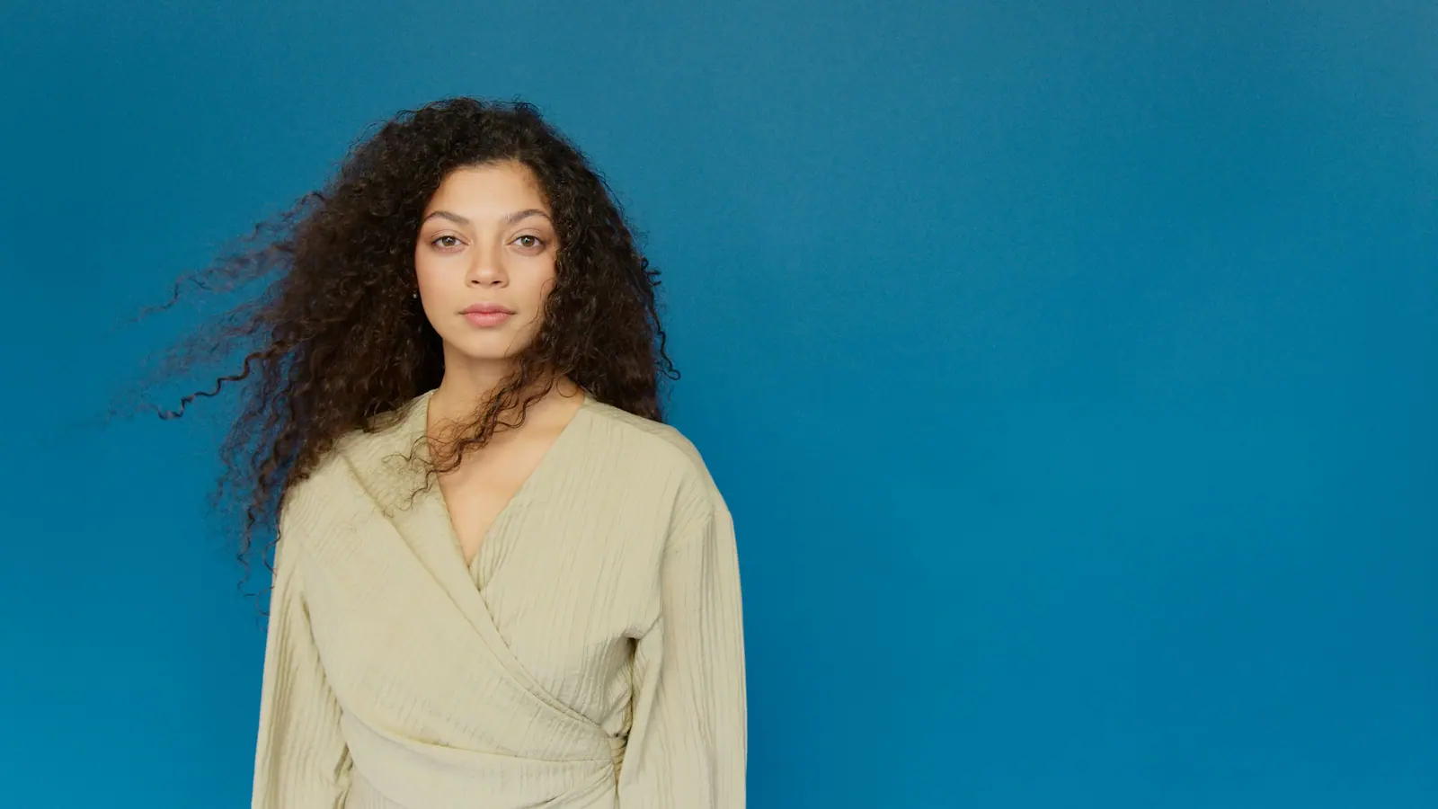 Woman with long dark curls in a sage wrap top against a deep blue wall