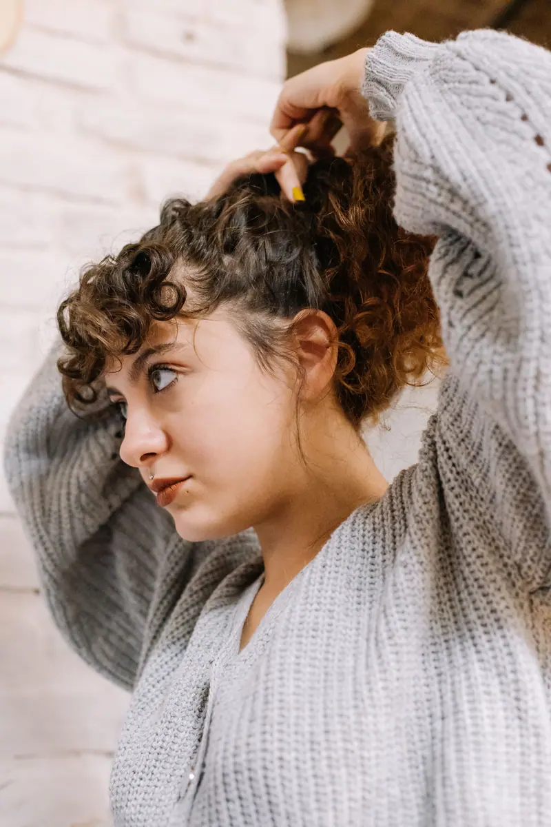 A young woman with curly hair ties her hair in a cozy indoor setting, wearing a knitted sweater.