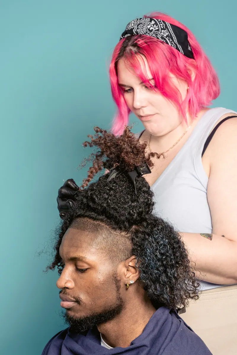 Hairdresser with vibrant pink hair styling curly hair in studio on blue background.
