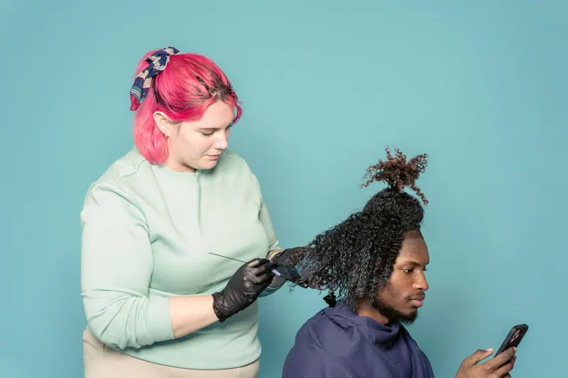 Professional hairdresser applying dye to client's curly hair while client uses smartphone.