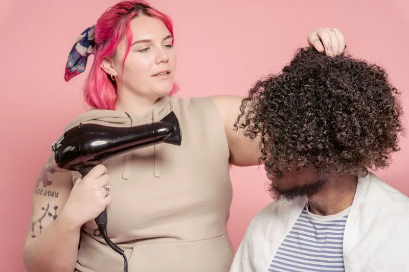 A hairdresser styling a man's curly hair using a hairdryer in a studio setting.