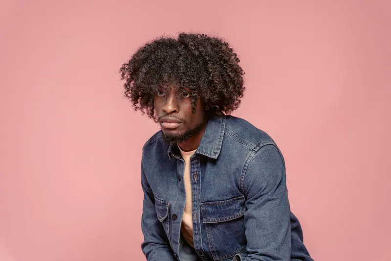 Confident young man with curly hair in a denim jacket against a pink background.