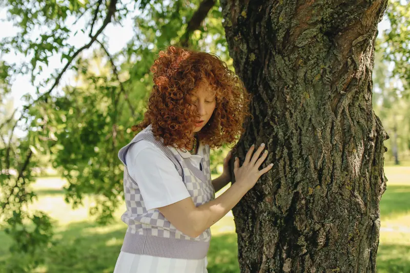 A woman with curly red hair gently touches a tree trunk in a sunny outdoor setting, surrounded by green foliage.