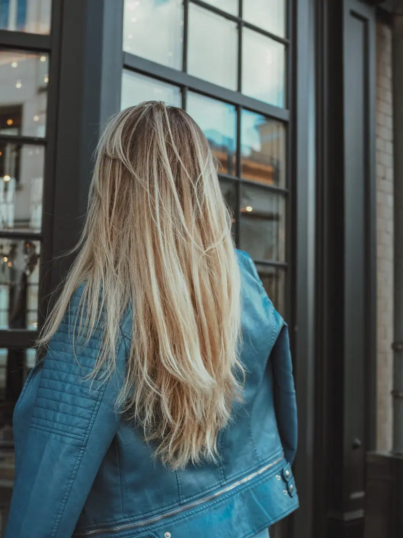 A woman with long blond hair in a blue jacket looks through a window in the city.