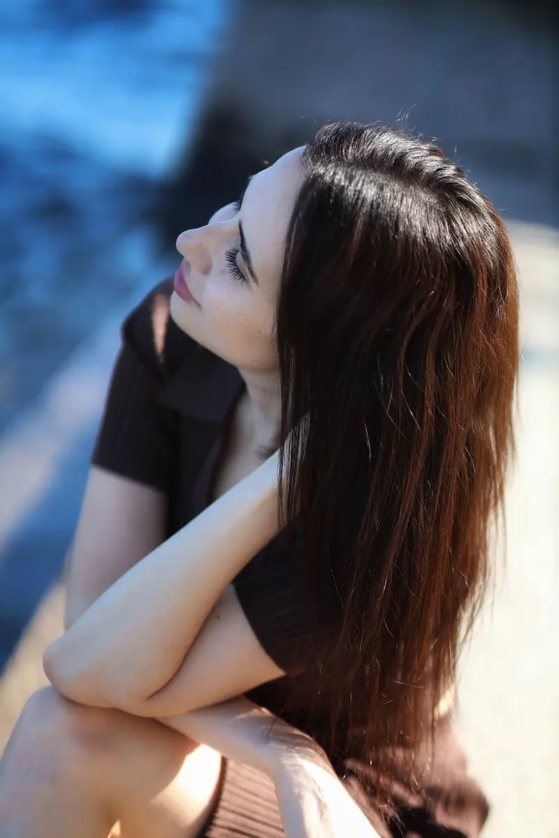 A woman with long hair seated outdoors, looking thoughtfully into the distance under natural light.