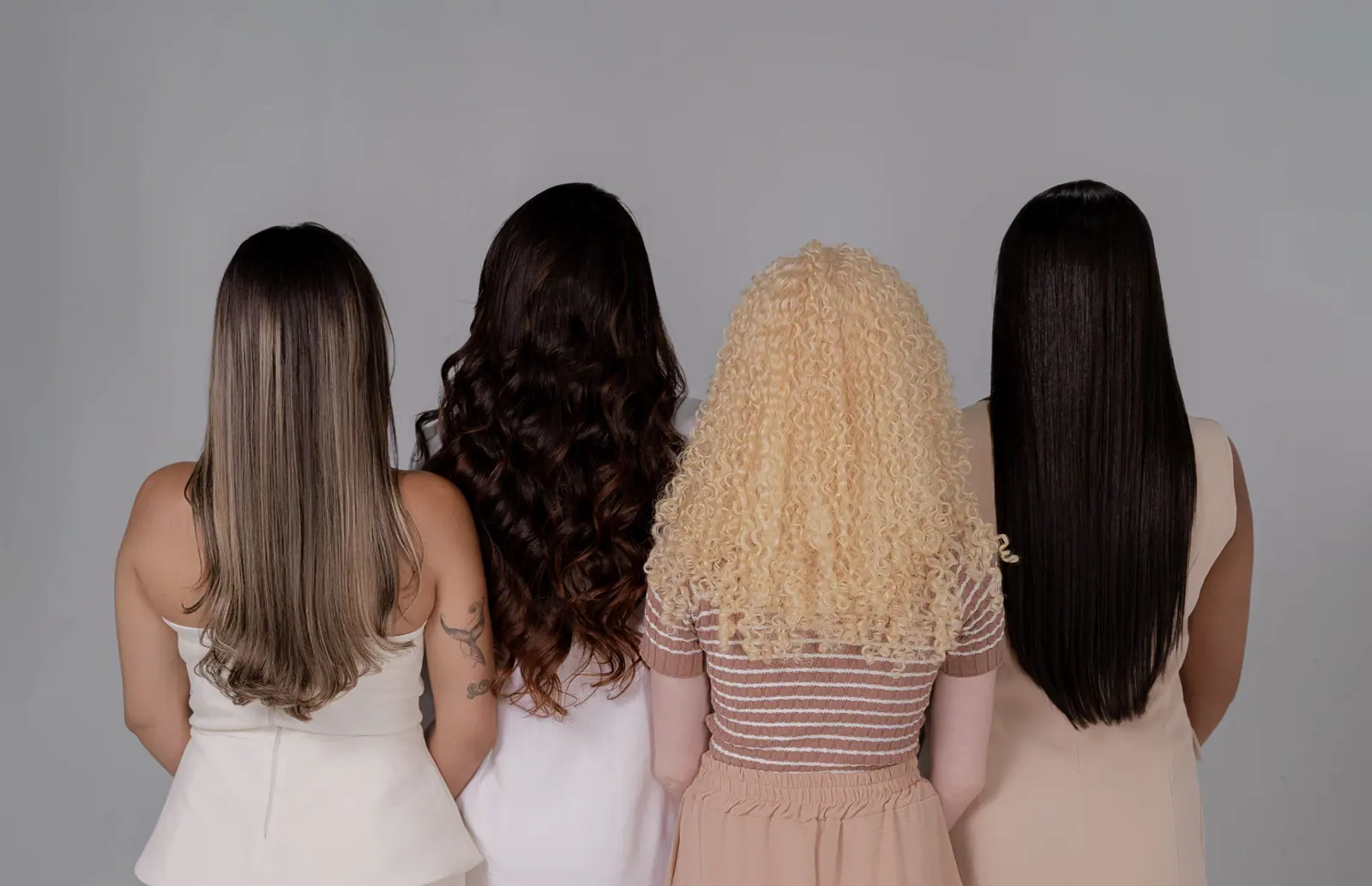 Four women from behind showing long brunette, curly, platinum and black hair against a greige wall