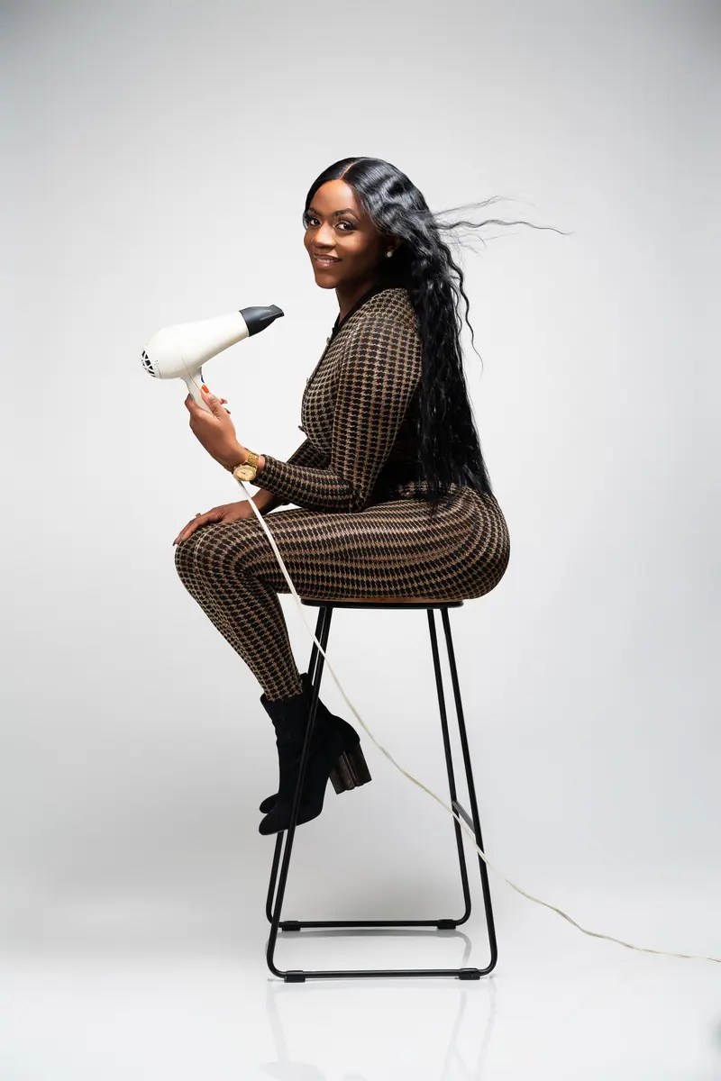 Elegant photo of a fashionable black woman with long hair using a blow dryer, poised indoors.