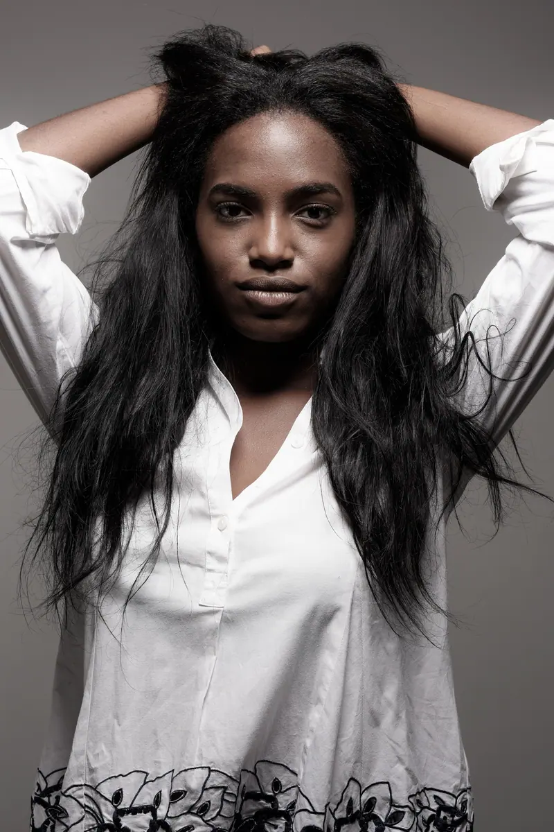 Sophisticated portrait of a woman with long hair in a white shirt, arms raised, against a neutral backdrop.