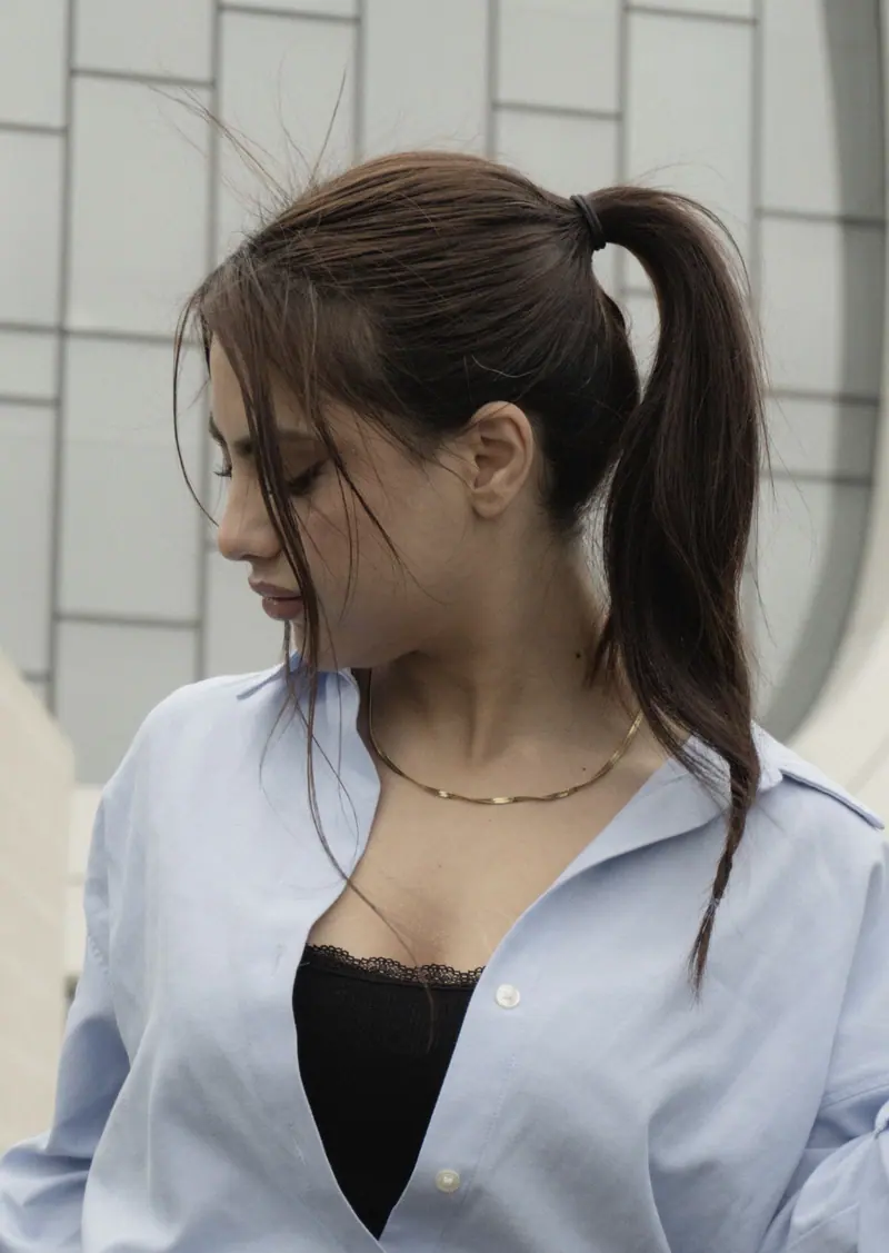 Portrait of a young woman with a ponytail wearing a blue shirt, captured in an outdoor setting.