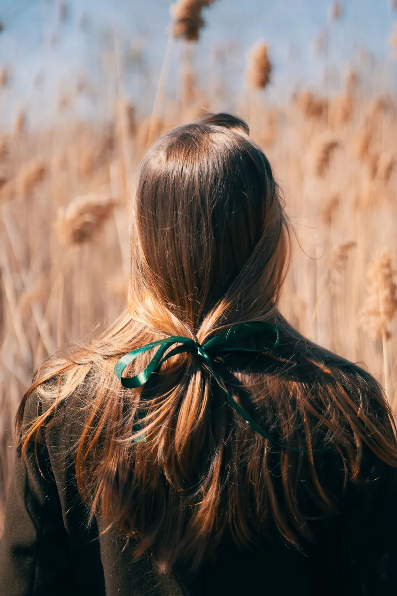 A woman stands in a wheat field with a green bow in her hair, surrounded by tall stalks under clear skies.