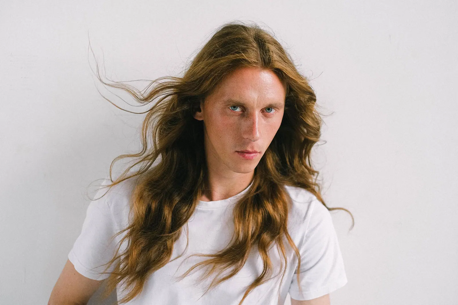 Woman with long, wind-tousled light-brown hair in a white T-shirt against a pale wall