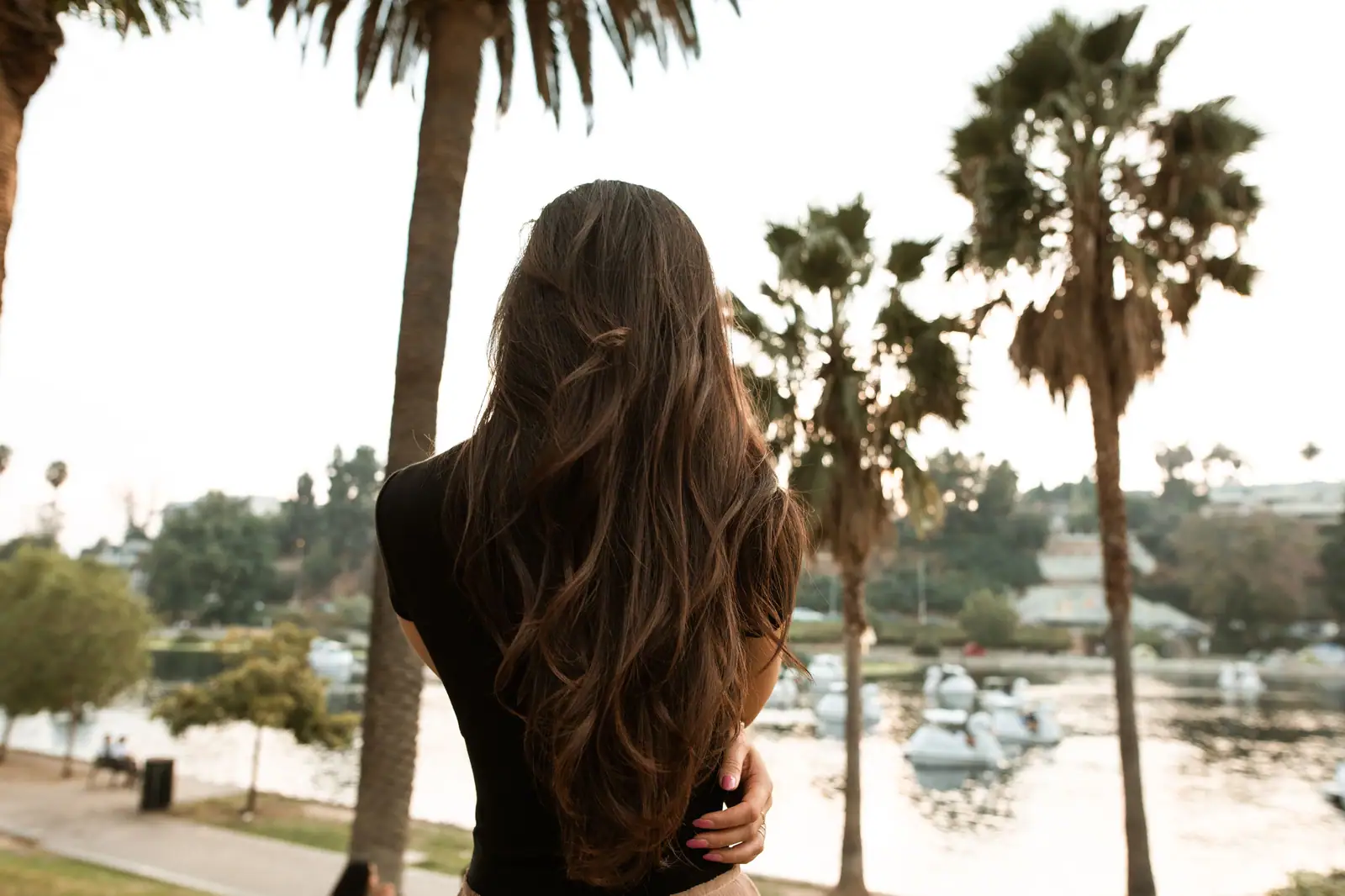 Long, sun-lightened brunette lengths seen from behind on a lakeshore lined with palms