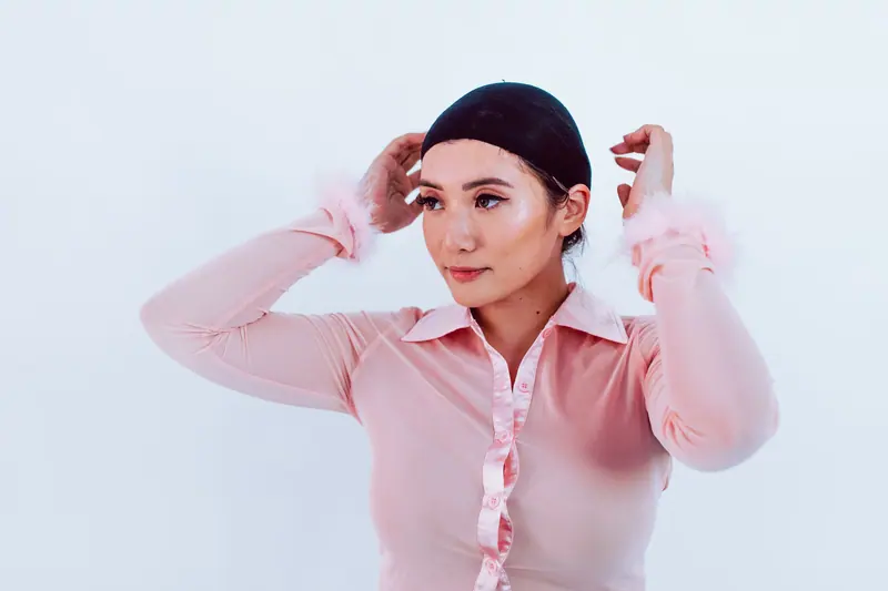 Woman in pink shirt adjusting her black hair cap indoors against white background.