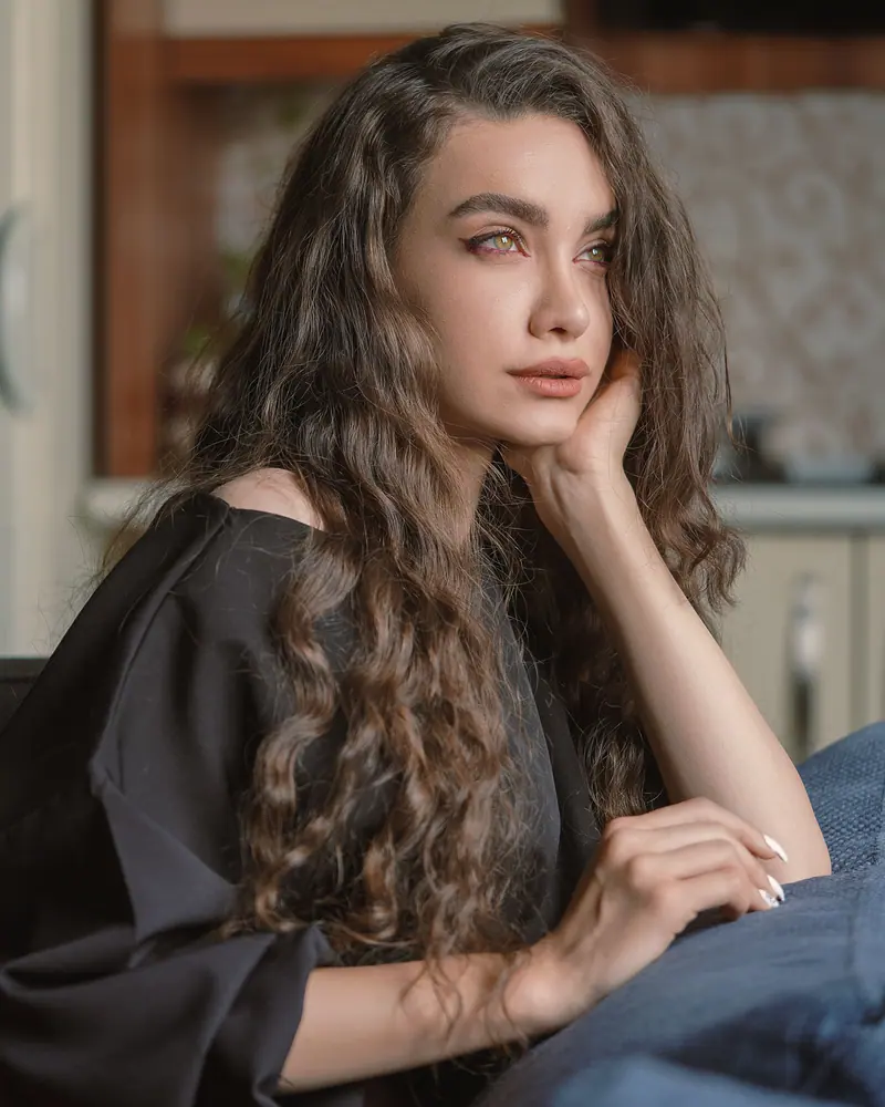 Elegant portrait of a woman with long wavy hair, lost in thought indoors.