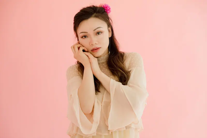 Stylish portrait of a woman posing with a pink flower in her hair against a soft pink background.