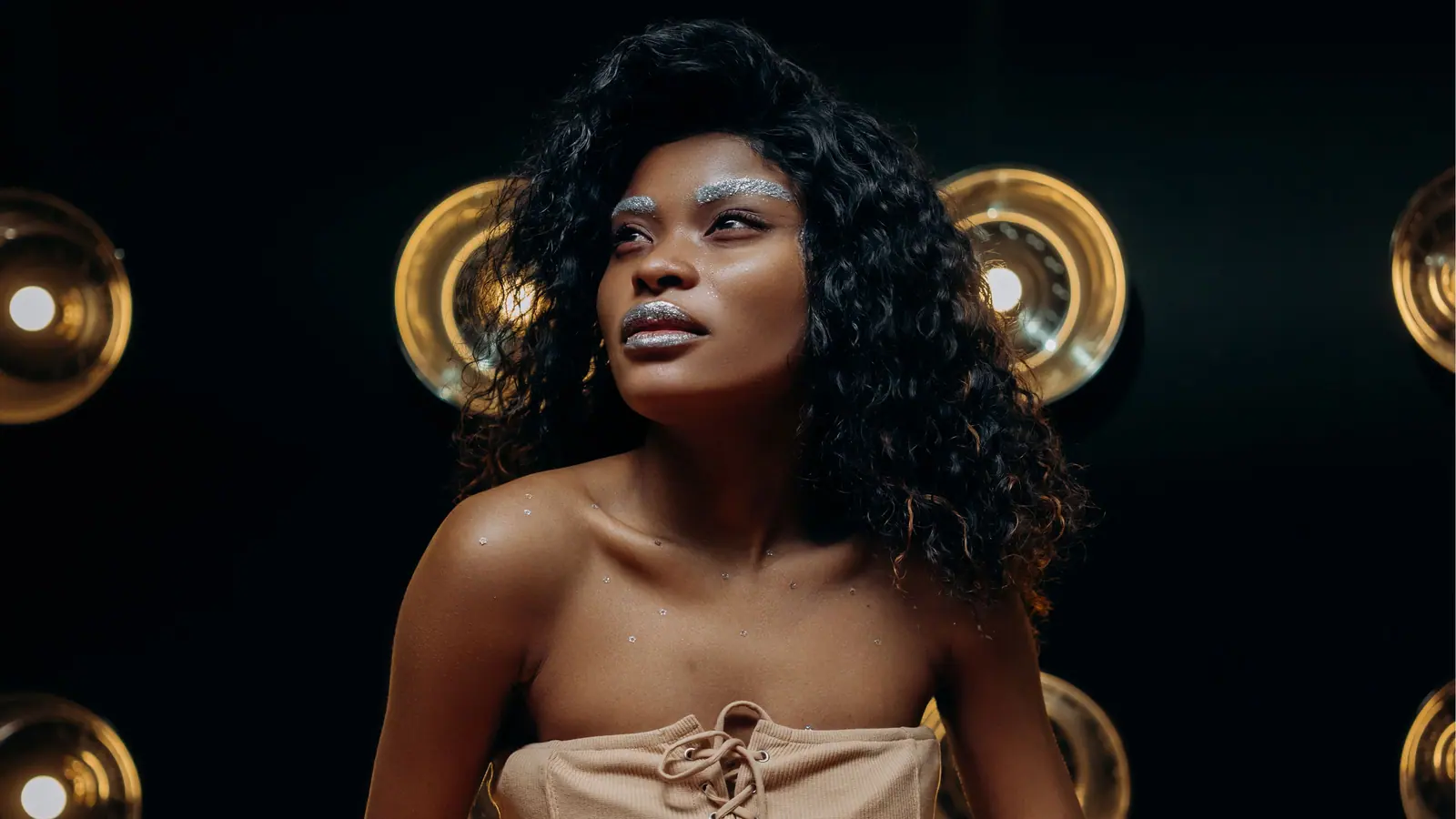 Woman with glossy black curls and silver glitter brows, lit against gold vanity bulbs