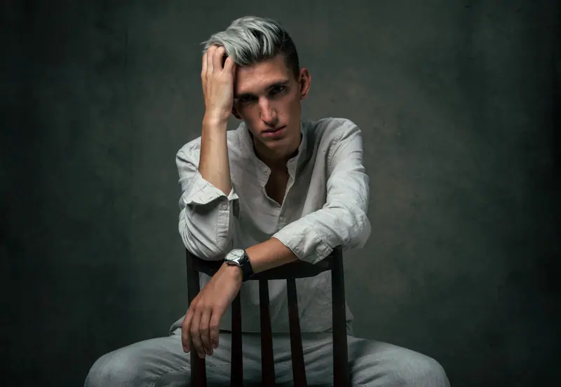 Man with a silver undercut leaning on a chair in a dark studio