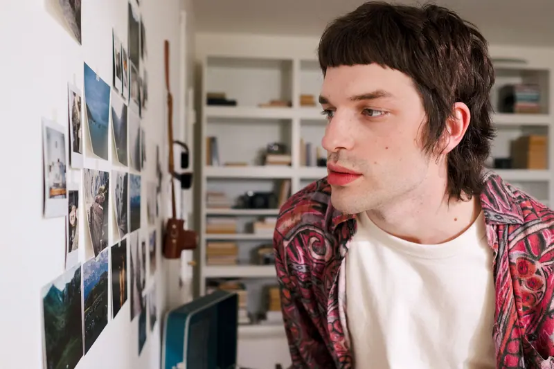 A young man closely observing a collection of pinned Polaroid photos on a wall indoors.