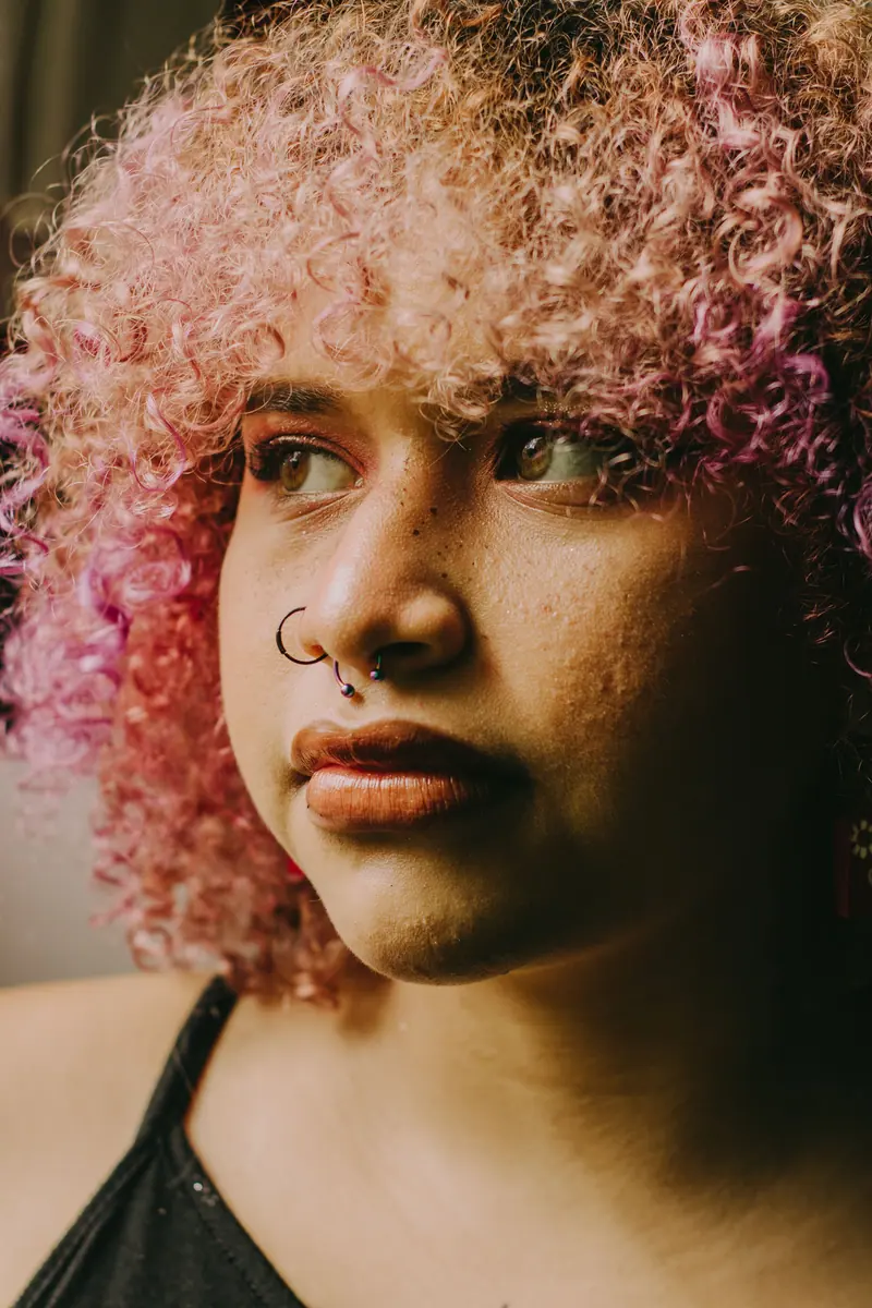 A striking close-up portrait of a woman with curly pink hair and nose piercings, looking thoughtful.