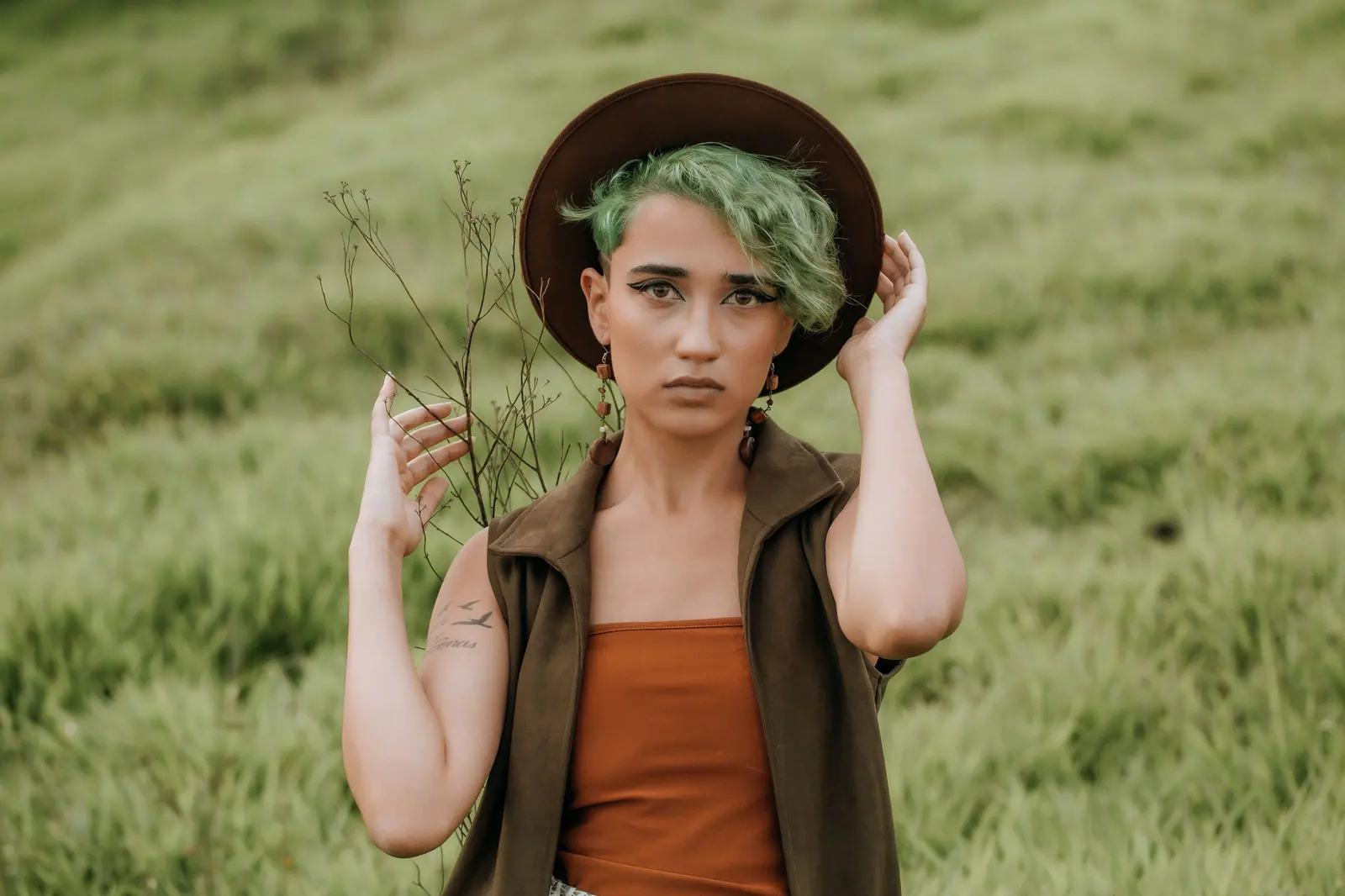 Person with a sage-green undercut and a brown felt hat standing in tall green grass