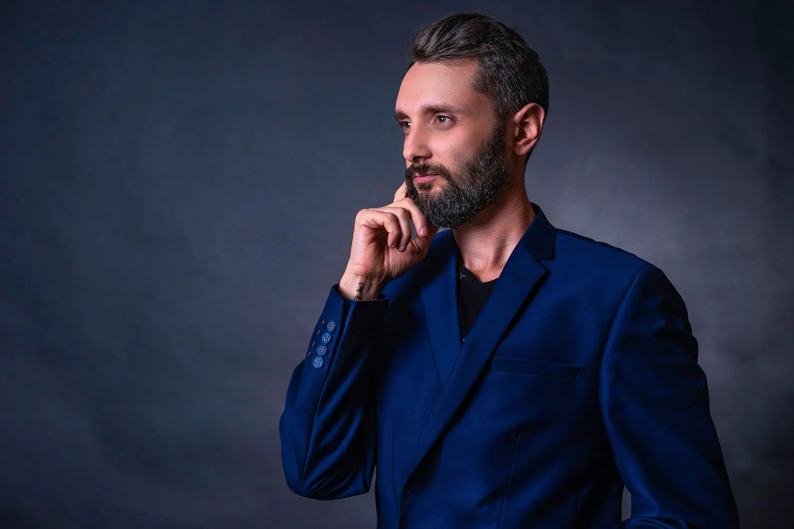 Confident businessman in a blue suit posing with hand on chin against a gray background.