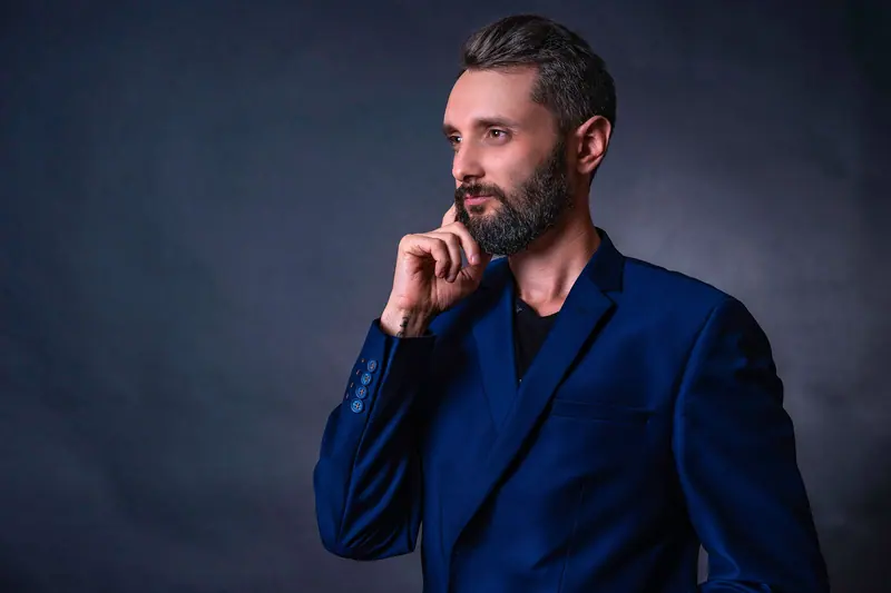 Confident businessman in a blue suit posing with hand on chin against a gray background.