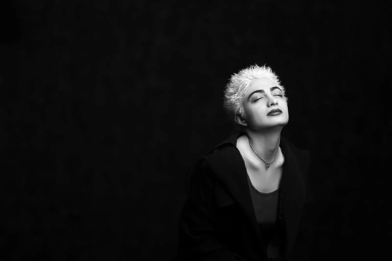 Black-and-white portrait of a person with a cropped platinum cut, head tilted back against a black backdrop