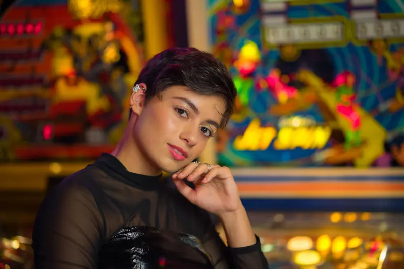 A young woman with short hair poses in a vibrant and colorful arcade setting.