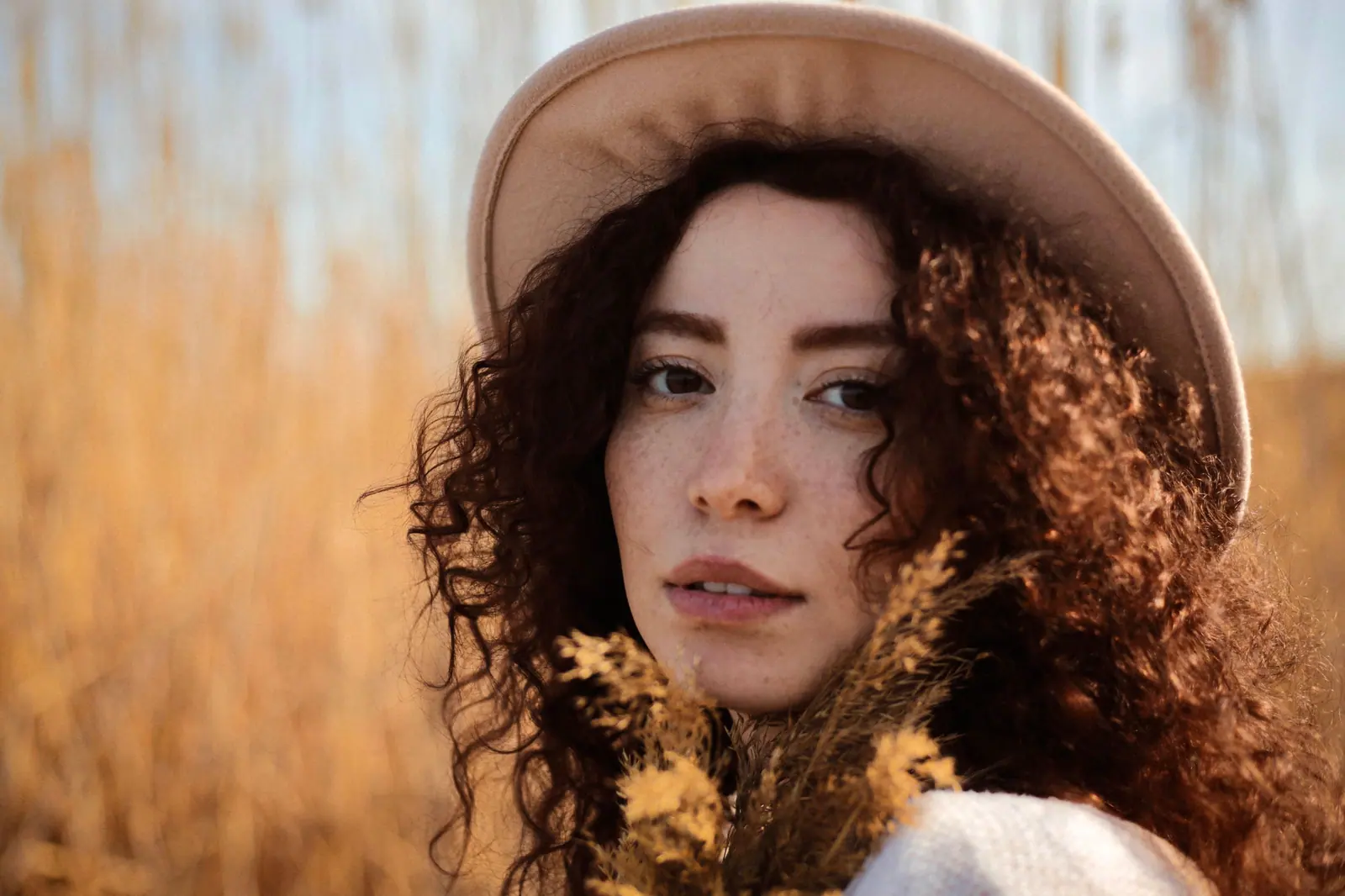 Woman with long dark curls and a cream felt hat holding dried grasses in warm golden light