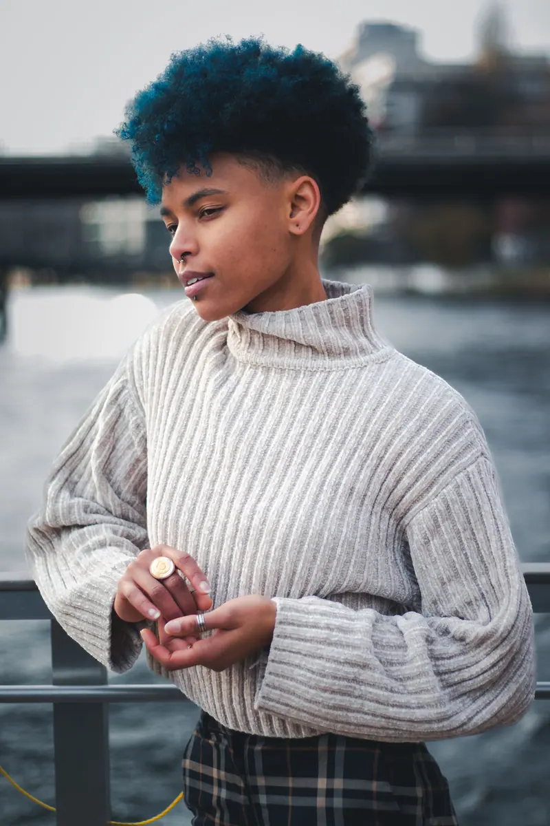 A stylish adult with blue hair and a septum piercing poses by a riverside in a cozy sweater.