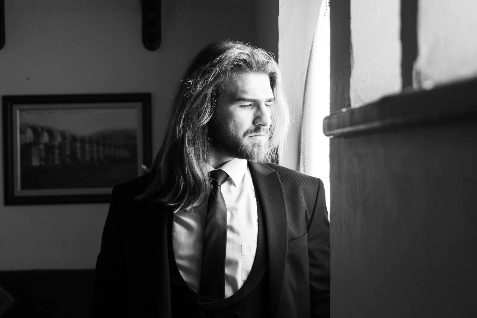 Black and white portrait of a groom with long hair in a tuxedo, looking out a window.