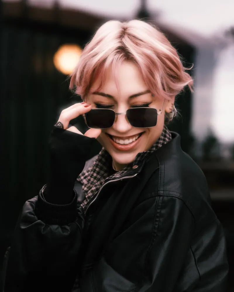 Chic woman with pink hair, smiling and wearing sunglasses outdoors.