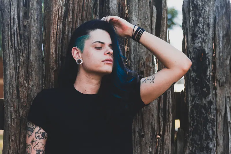 A trendy young man with tattoos and blue hair posing against a wooden fence, exuding confidence and individuality.