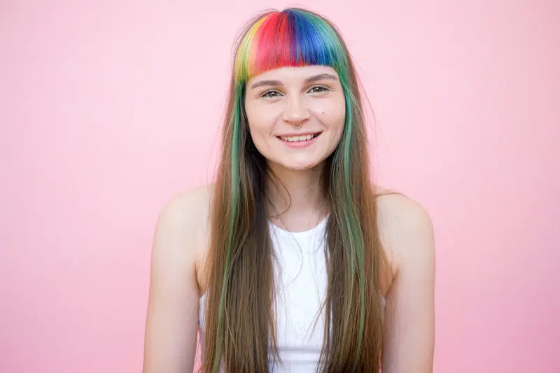 Rainbow-dyed fringe and long hair against a pink wall