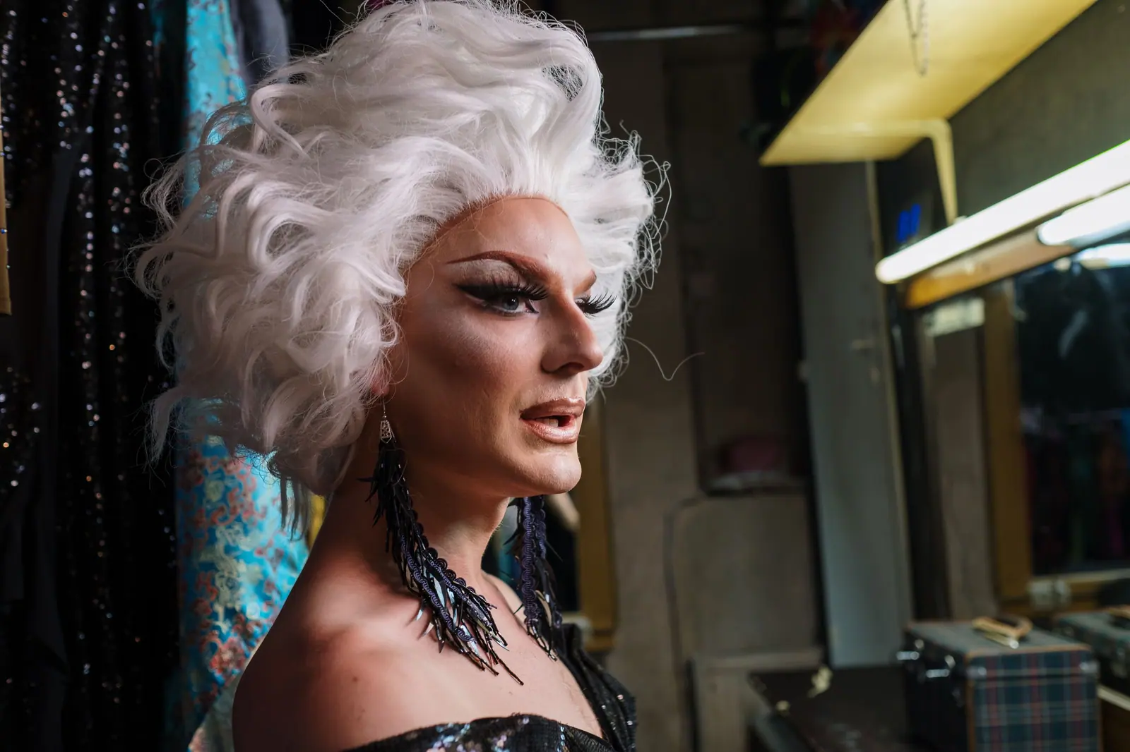 Drag performer with a sculpted platinum wig and long jeweled earrings in a warm, cluttered dressing room